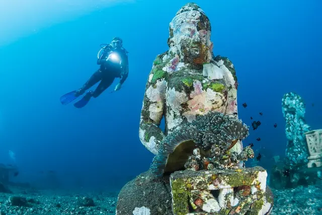 An underwater Buddha with a diver holding a light behind it
